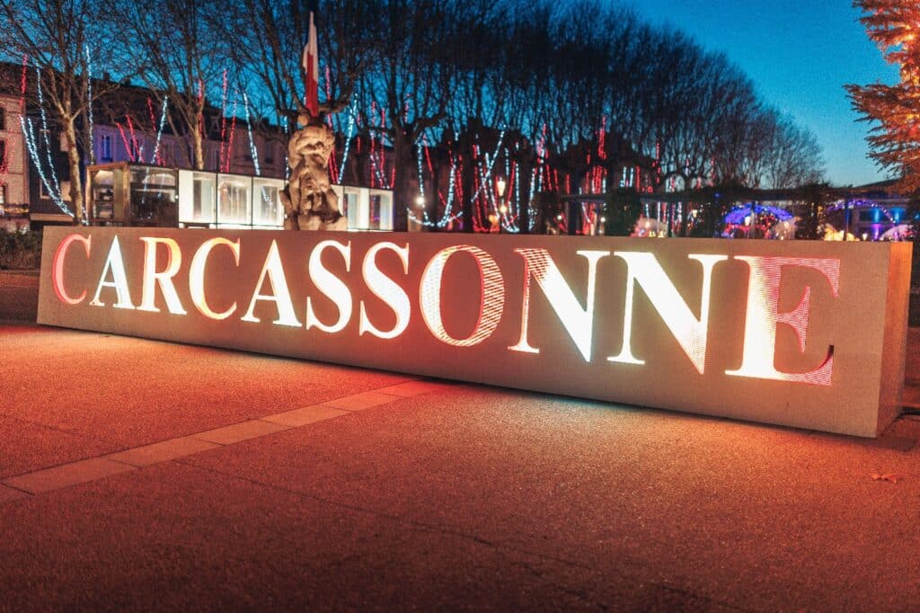 Enseigne lumineuse Carcassonne en soirée avec arbres décorés.