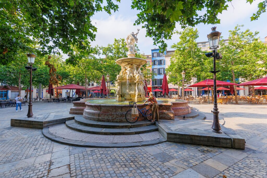 Fontaine sur une place avec terrasses ombragées.